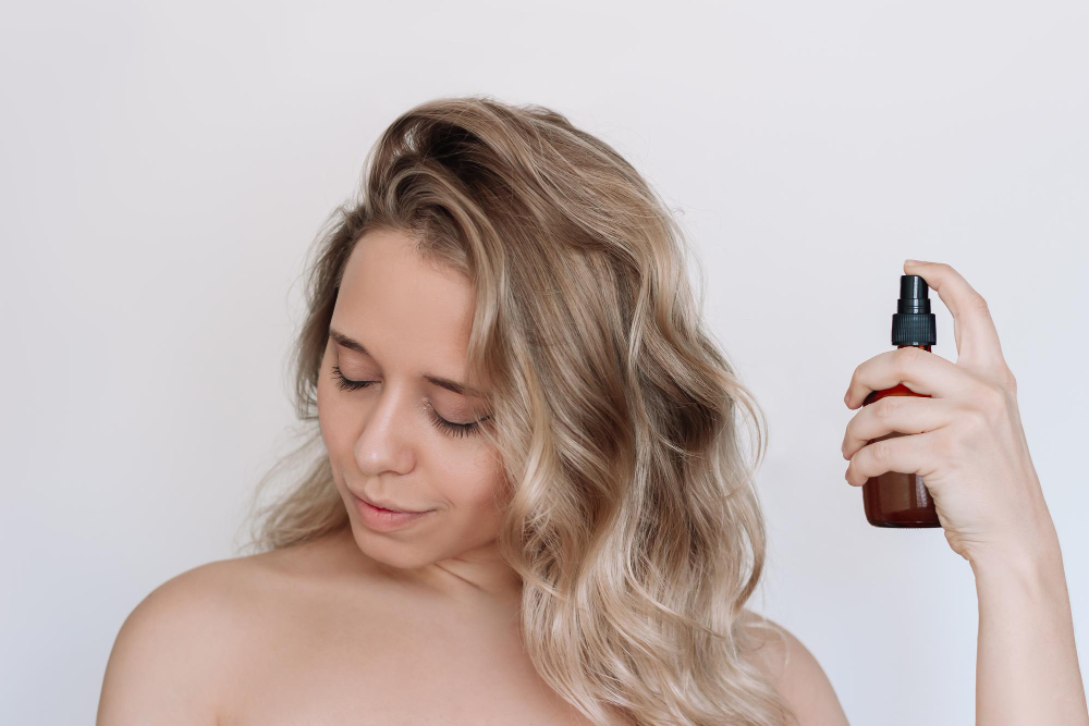 Kobieta z falowanymi blond-platynowymi włosami, aplikująca serum do włosów w sprayu, na białym tle.