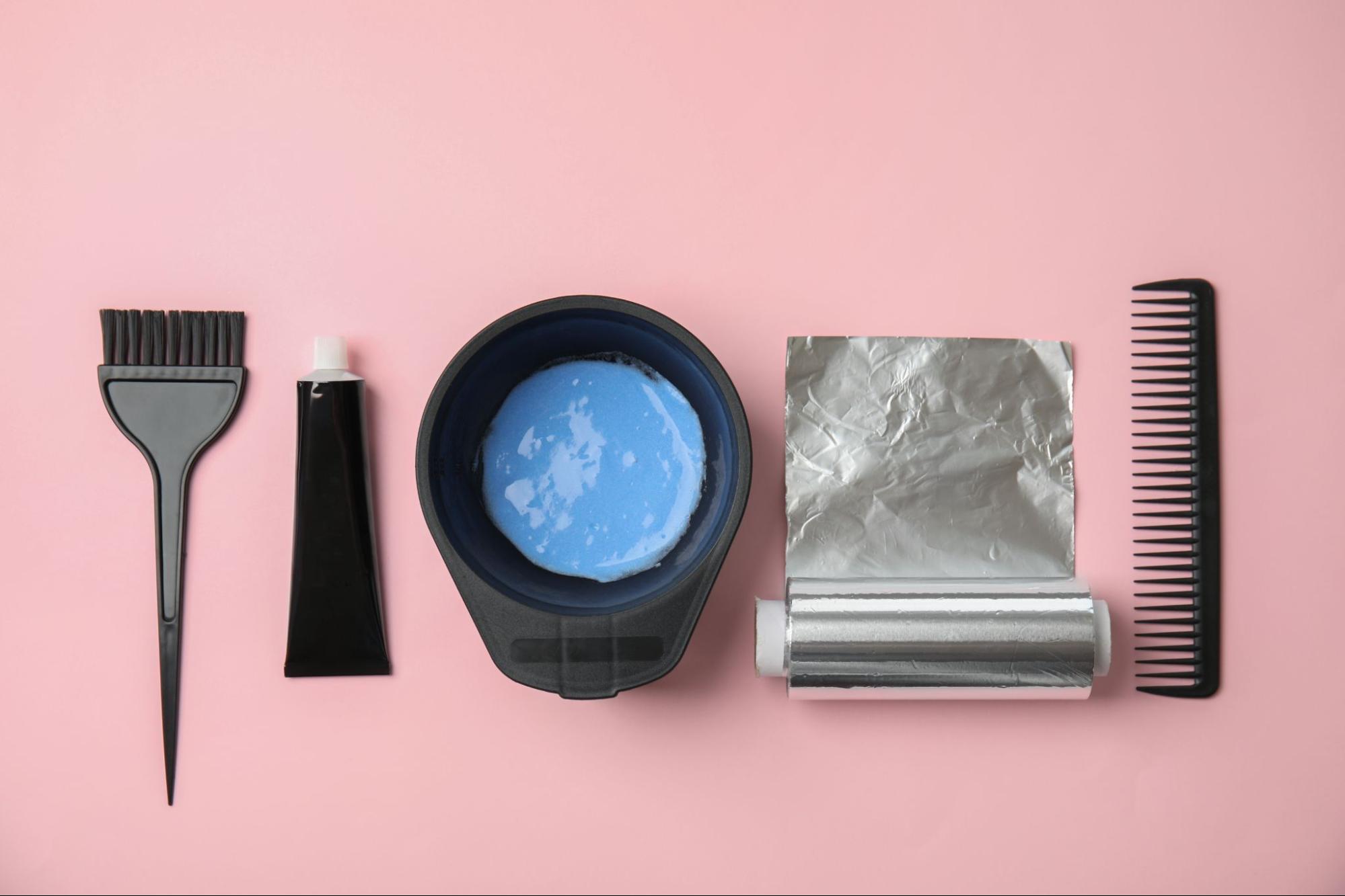 Pędzelek, aktywator, toner, folia i grzebień rozłożone obok siebie na różowym tle. 